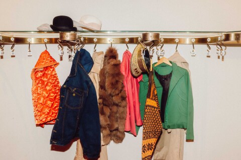 En rekke fargerike jakker og kåper henger på en messingfarget garderobestang med nummererte kroker. Plaggene inkluderer blant annet en oransje vattert vest, en denimjakke, en brun pelsvest, en rød kåpe, en grønn blazer og et skjerf med gult og brunt mønster. Øverst på stangen ligger flere hatter, inkludert en svart bowlerhatt og en lys stråhatt.