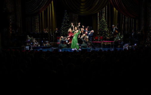 Kvinne synger, mens en gruppe artister står rundt og strekker armene mot henne. Alle er festkledd