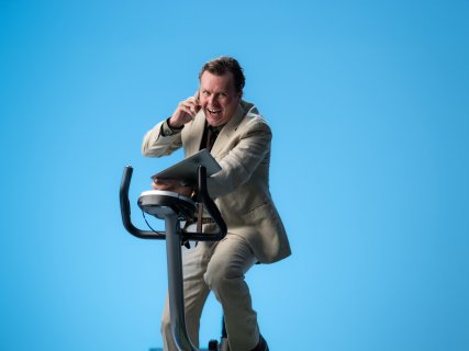 Mann med oppkavet uttrykk på trimsykkel mot blå bakgrunn, med frakk, PC og mobil i hendene, skjorten er delvis åpen og uordnet.