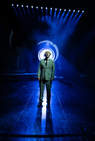 Mann i dress på mørk scene med blått lys. Et stort øye i bakgrunnen.