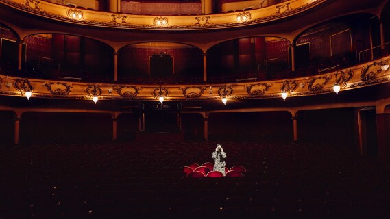 En kvinne i mønstret antrekk står alene blant røde fløyelsseter i en stor, mørklagt teatersal. Hun ser opp og holder hendene mot ansiktet, som om hun beskytter øynene mot lys eller speider mot scenen. Rundt henne er salen tom, mens lysekroner og gulldekorasjoner lyser opp de øvre balkongene.