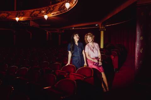 To kvinner sitter i en mørklagt teatersal med røde fløyelsseter og snakker og ler sammen. Den ene har på seg en mørkeblå kjole, den andre en rosa topp og skjørt, og holder et glass vin. De sitter avslappet ved siden av hverandre foran scenen, omgitt av teatrets klassiske, elegante interiør.