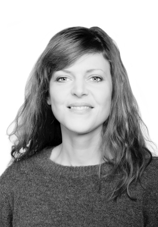 Sort/hvitt-bilde av kvinne i 30-årsalderen med langt brunt hår, som smiler til kamera. Hun har på en mørk strikkegenser.