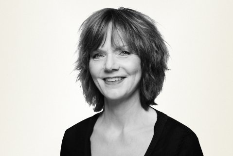 Svart-hvitt-bilde av kvinne i 60-årene med kort, mørkt hår med lugg og svart genser som ser skrått inn i kamera og smiler. Lys bakgrunn.