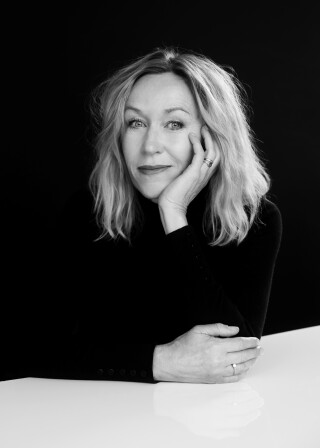 Svart-hvitt-portrett av kvinne i 50-årene med blondt, skulderlangt hår og svart genser. Hun hviler ansiktet i venstre hånd og albuene på et bord.